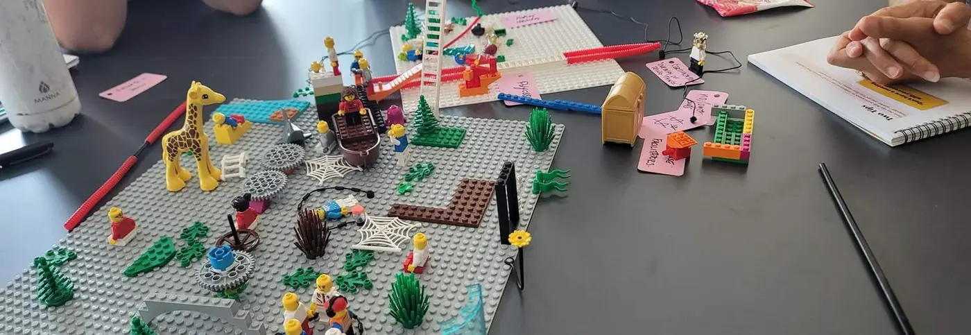 A shared LEGO Serious Play model on the table, part way through a systems build, with handwritten meaning cards around it
